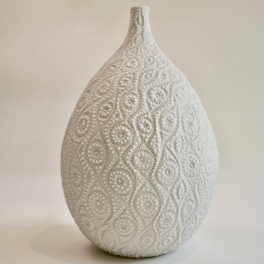 A teardrop-shaped ceramic vase with a textured, raised floral lace-like pattern in a warm off-white or light beige tone, set against a plain, neutral background.
